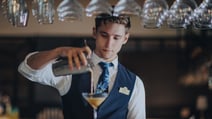 A stylish barman in a suit serves a drink into a glass at the bar of the Runnymede on Thames hotel.